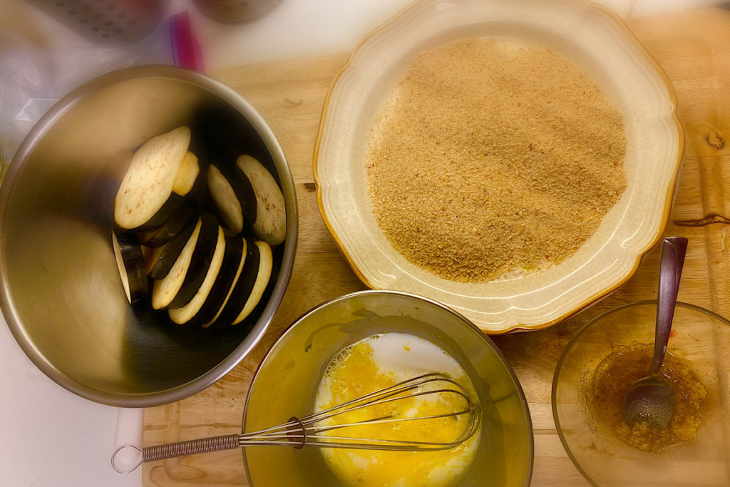 A bowl with eggplants marinate, a plate with bread crumbs, small bowl with egg and milk, other small bowl with garlic , olive oil and salt to seasoning the eggplants.